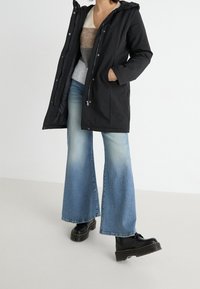 Giacca nera impermeabile con cappuccio, dotata di chiusura con zip e bottoni a pressione; abbinata a jeans svasati azzurri chiari e stivaletti neri con tacco alto.