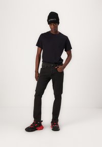 Camiseta negra, jeans ajustados negros con un desgastado en la rodilla y zapatillas negras con acentos rojos. Llevado con un gorro de lana negro.