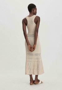 Abito lungo in maglia beige con corpetto aderente, schiena scoperta e orlo svasato con motivo in pizzo. Il modello indossa sandali flat.