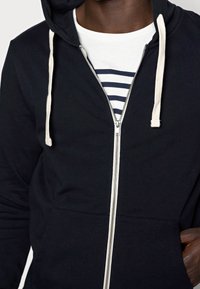 Černá mikina na zip s texturovanou textilií, se zvýrazněnou krémovou šňůrkou a zipem. Na přední straně jsou dvě kapsy.