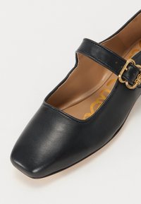 Pantofi Mary Jane din piele neagră, cu vârf pătrat, detaliu cu cataramă pe lateral și o branț bej cu un model de logo galben.