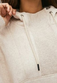 Sudadera beige clara con una textura suave, que presenta un panel de diseño acanalado, cordones ajustables con puntas negras y una capucha acogedora.