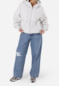 Světle šedá mikina na zip s kapucí a bílými šňůrkami, spárovaná s volnými modrými džíny s roztrženou záplatou a rovnými nohavicemi.