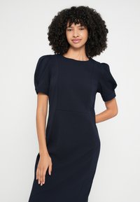 Tumši zila kleita ar strukturētu siluetu, īsām pūkainām piedurknēm un šuvju detaļām, izgatavota no teksturēta, elastīga auduma.