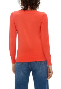 Vrouw met krullend haar, gekleed in een feloranje lange mouwtrui en blauwe spijkerbroek, van achteren gezien op een witte achtergrond.