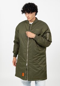 Jonge man die een te grote olijfgroene bomberjack met ribgebreide manchetten en een oranje patch bij de zoom dichtritst, terwijl hij lichte jeans draagt tegen een witte achtergrond.