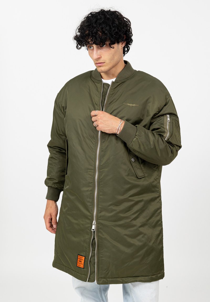 Jonge man die een te grote olijfgroene bomberjack met ribgebreide manchetten en een oranje patch bij de zoom dichtritst, terwijl hij lichte jeans draagt tegen een witte achtergrond.