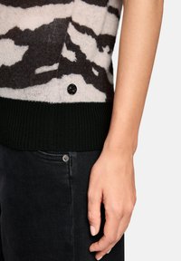 Gestrickter Pullover mit einem schwarz-weißen abstrakten Muster, einem dunkelgrünen, gerippten Saum und schwarzen Knopfdetails nahe dem unteren Rand.