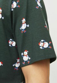 Grøn T-shirt med korte ærmer, der har gentagne mønstre af tegneserie-snefnug med julesokker, i røde og blå nuancer. Blødt bomuldsstof.