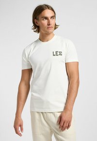 Camiseta blanca de manga corta hecha de algodón. Presenta un logo verde "LEE" en el lado izquierdo del pecho. Textura suave y cuello redondo clásico.