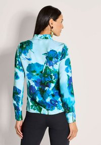 Woman with long dark hair wearing a light blue blouse with large blue and green floral print, paired with black pants, shown from the back.