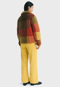 Chaqueta de forro polar texturizada y multicolor con un patrón de cuadros en rojos, marrones y amarillos; combinada con pantalones de pana amarillos.