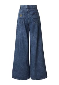 Platus tamsiai mėlynas džinsinių audinių kelnių modelis su aukštu liemeniu, dviem užpakalinėmis kišenėmis ir logotipo ženkliuku šalia dešinės kišenės. Tekstūruota medžiaga.