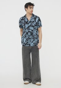Camicetta a maniche corte con sfondo nero e motivo di foglie tropicali blu chiaro. Abbinata a pantaloni in denim grigi a gamba larga.