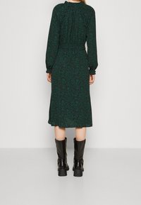 Långärmad, mörkgrön klänning med blommönster, med en rynkad halsringning och elastiska ärmslut, kombinerad med knähöga svarta stövlar.