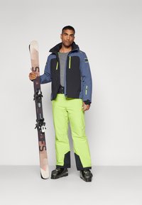 Equipo de esquí con una chaqueta azul y negra con detalles verdes, pantalones verde neón y botas de esquí; sosteniendo un par de esquís.