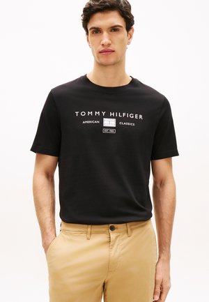Junger Mann trägt ein schwarzes Tommy Hilfiger T-Shirt mit weißem Logo-Schriftzug und beigen Hosen, steht mit einer Hand in der Tasche.