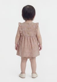 Kleinkind mit kurzen dunklen Haaren, das ein rosa Blumenkleid mit Knöpfen und gerafften Ärmeln sowie beige Schuhe trägt, steht mit dem Rücken zum Betrachter auf weißem Hintergrund.