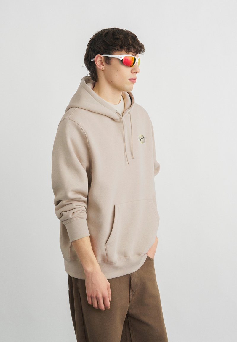 Giovane uomo con felpa con cappuccio beige, pantaloni marroni e occhiali da sole sportivi bianchi con lenti riflettenti rosse, in piedi davanti a uno sfondo chiaro uniforme.