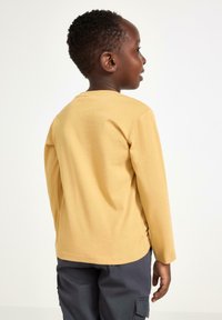 Langærmet gul bomulds T-shirt med rund hals, glat tekstur og afslappet pasform. Set fra ryggen, parret med mørke bukser.