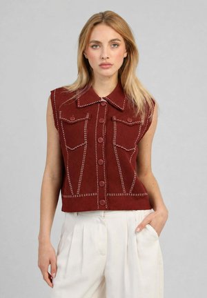 Femme blonde portant un gilet sans manches rouge foncé boutonné avec des coutures blanches et un pantalon blanc plissé taille haute, une main dans la poche.