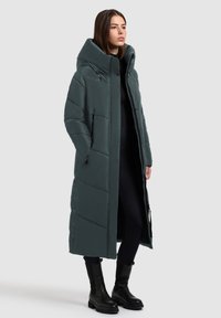 Duga, podložena jakna tamnozelene boje s visokim ovratnikom, prednjim zatvaračem, bočnim džepovima i dugmadima na dugmetu uz rub. Glatka, sjajna tekstura.