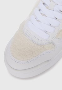 Weißer Sneaker mit einem flauschigen beigen Textil-Obermaterial, glatten Lederakzenten, flachen weißen Schnürsenkeln und einer dicken, strukturierten Gummisohle.