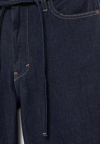 Mørkeblå denimjeans med synlig forlomme, bæltestrop og matchende denim bælte, der hænger ned foran.