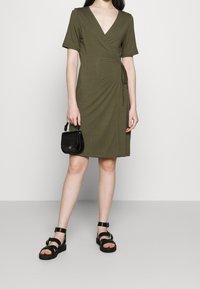 Robe portefeuille vert olive avec un décolleté en V profond, des manches courtes et un détail de lien sur le côté, associée à des sandales noires à lanières et un petit sac noir.