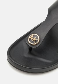 Infradito neri con una superficie di appoggio testurizzata, fascia liscia e un accento circolare in oro con il logo MK sulla fascia. Design a suola piatta.