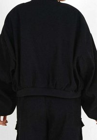 Persoon in een losse zwarte sweatshirt met een gerimpelde zoom en bijpassende zwarte broek, gezien vanuit de achterkant met de armen ontspannen langs het lichaam.