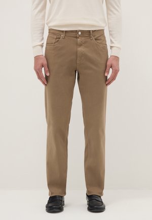 Man draagt beige rechte broek, zwarte schoenen en een crème gekleurde lange mouwen blouse, staand tegen een effen lichte achtergrond.