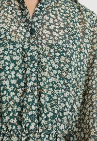 Pohjavihreä kukallinen paitamekko, jossa läpikuultava kangas. Mekossa on pieniä valkoisia kukkia ja oransseja yksityiskohtia, vyötäröllä laskokset ja V-pääntie.