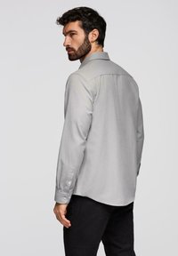 Homme aux cheveux foncés et à la barbe, portant une chemise à manches longues gris clair et un pantalon noir, légèrement tourné sur le côté contre un fond uni.