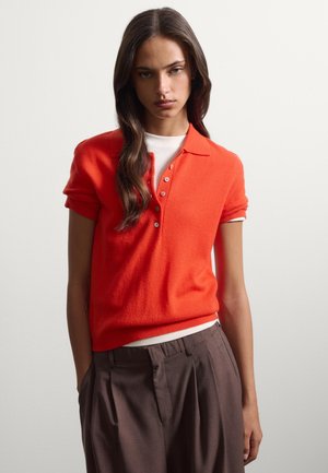 Joven mujer con cabello largo y oscuro que lleva una camiseta polo de manga corta de color naranja brillante sobre una camiseta blanca y pantalones de tiro alto de color marrón oscuro.
