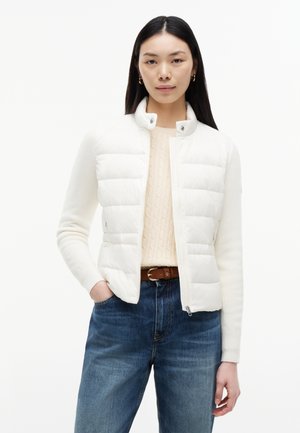 Frau mit langen schwarzen Haaren, die eine weiße gesteppte Jacke, einen cremefarbenen Zopfpullover, blaue Jeans und einen braunen Gürtel trägt, steht mit einer Hand in der Tasche.