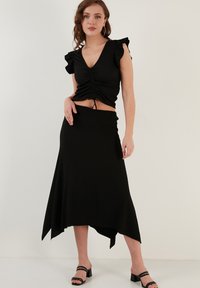 Juoda banguota palaidinė su raukiniais ir raišteliu, derinama su juoda sijonu aukštesne priekyje ir žemesne gale. Modelis vilki juodus sandalus su juostelėmis.