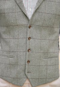 Szara kamizelka w jodełkę z brązowym wzorem w kratę, wyposażona w pięć okrągłych guzików, dwie boczne kieszenie oraz klapę z wycięciem. Tkanina o fakturze.