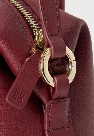 Gros plan d'un sac à main en cuir bordeaux avec une fermeture éclair dorée et une boucle circulaire dorée.