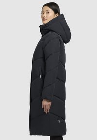 Sort lang pufferjakke med vandret quiltning, høj krave og fastgjort hætte. Har sidelommer og et logo på kanten. Glat tekstur.