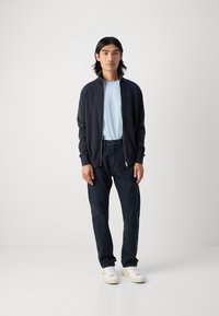 Mornarica zip-up kardigan preko svijetloplavog majice, uparen s tamnim hlačama i bijelim tenisicama. Model stoji protiv jednostavne pozadine.