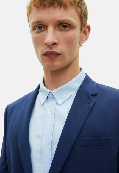 Ein marineblauer Blazer aus strukturiertem Stoff mit einer einzigen Brusttasche, kombiniert mit einem hellblauen Hemd mit Kragen und Knopfverschluss.
