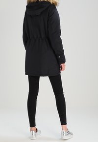 Person klädd i en svart vinterparka med pälshuva, svarta leggings och silverfärgade sneakers, stående med ryggen mot kameran mot en enfärgad bakgrund.