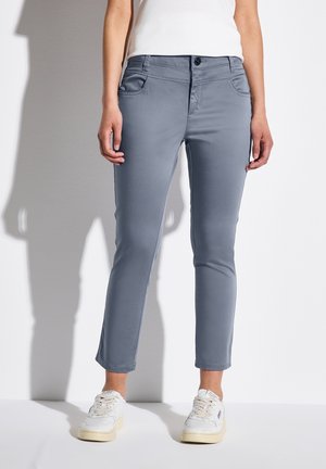 Vrouw die slanke grijze broek en witte sneakers draagt, staand tegen een witte muur met een schaduw naar links.