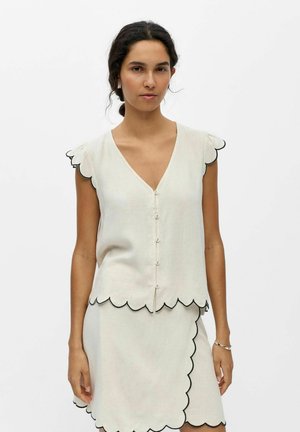 Femme portant un chemisier crème sans manches et une jupe assortie aux bords festonnés soulignés en noir, debout sur fond blanc.