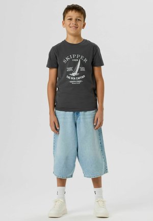 Garçon debout, portant un t-shirt gris foncé "Skipper" avec un motif de voilier, un short en jean bleu clair long, des chaussettes blanches et des baskets blanches.