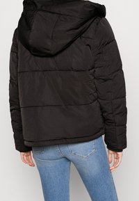 Persona che indossa una giacca a vento nera con cappuccio e jeans azzurri chiari, vista da dietro su uno sfondo neutro.