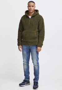 Olive green fleece pullover hoodie with a front pocket, half-zip collar, and drawstrings. Paired with blue denim jeans and black sneakers.