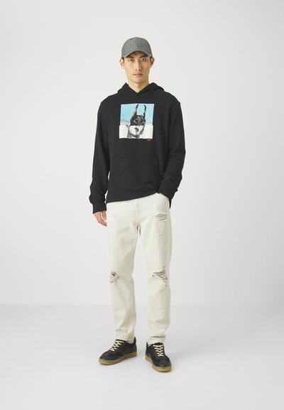 Hoodie noire avec un graphique sur le devant représentant un visage et du texte, associé à un jean blanc déchiré et des chaussures noires avec des lacets jaunes. Casquette grise portée.