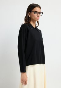 Schwarzer Strickpullover mit rundem Kragen und Perlendetails. Lange Ärmel und eine lockere Passform, kombiniert mit einem hellen Rock.